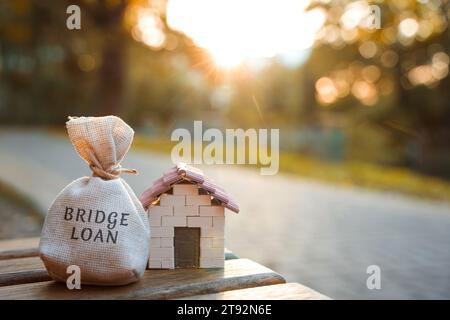 Bridge Loan Concept - è un prestito a breve termine garantito da immobili. Borsa di denaro e casa in miniatura nel parco alla luce del tramonto. Immobili e finan Foto Stock