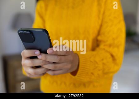 Sezione centrale della donna birazziale in maglione giallo che usa lo smartphone a casa Foto Stock