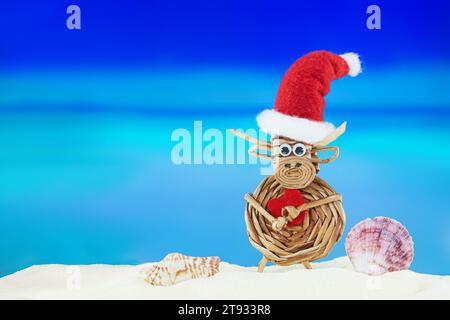 Mucca tessuta giocattolo con cappello di Babbo Natale con cuore e conchiglie sulla spiaggia sabbiosa con mare. Natale, Capodanno, San Valentino. Paesi caldi. Copia spazio Foto Stock