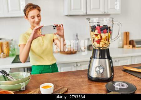 donna felice con lo smartphone che scatta foto al frullatore elettrico con frutta tritata, ricetta vegetariana Foto Stock