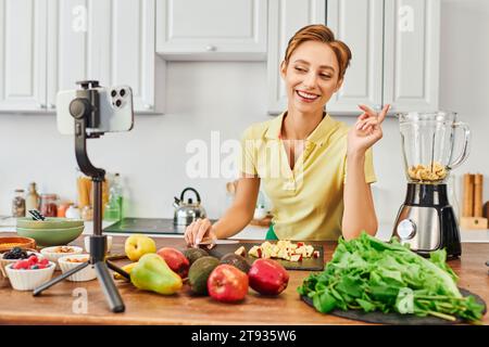 allegra donna vegetariana che guarda lo smartphone sul treppiede vicino alla frutta e al frullatore elettrico, vlog Foto Stock