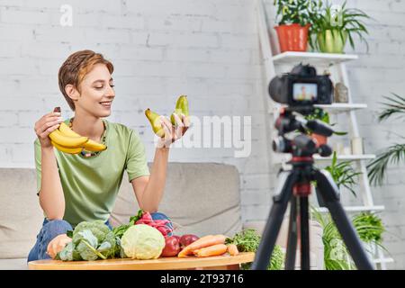 donna vegetariana con banane mature sorridente durante il video blog vicino a frutta e verdura fresca Foto Stock