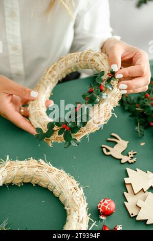 Da sotto il raccolto anonimo femminile che decora e tiene la corona di Natale con rami verdi con bacche rosse, sfondo verde accanto al diff della corona Foto Stock