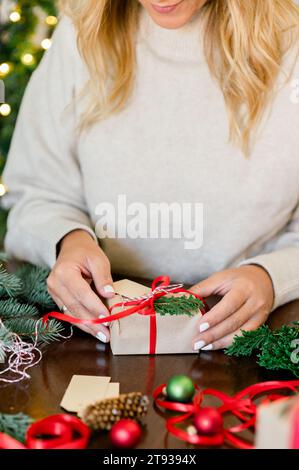 Raccolto di donna irriconoscibile in maglione che regge il regalo di Natale decorato con nastro rosso e abete in mano sul tavolo, con luci natalizie sullo sfondo Foto Stock