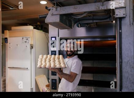 Ritratto di un giovane panettiere arabo con una maglietta bianca e un berretto rosso che porta un vassoio con una moltitudine di piccole palle di impasto sorridenti in un panificio con la stufa Foto Stock