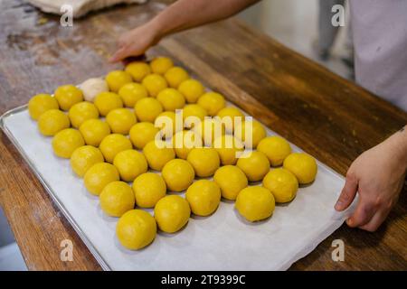 Ritratto di una moltitudine di piccole palline di pasta gialla su un vassoio su un tavolo mentre le mani di un fornaio la sollevano in un panificio Foto Stock