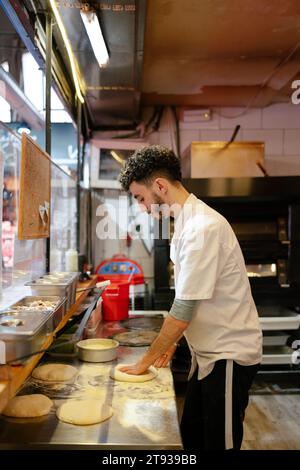 Ritratto di un giovane pizzaiolo arabo in grembiule bianco che prepara la pizza manualmente sul tavolo di una pizzeria Foto Stock