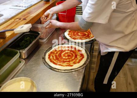 Scatto dettagliato delle mani di un uomo della pizza irriconoscibile che prende il prosciutto da un contenitore da aggiungere alla pizza in una pizzeria Foto Stock