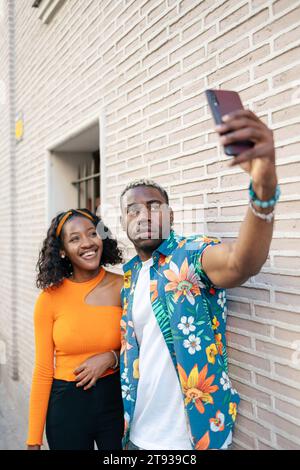 Giovane coppia nera che indossa abiti casual e colorati scattando un selfie e sorridendo mentre cammina per la città Foto Stock