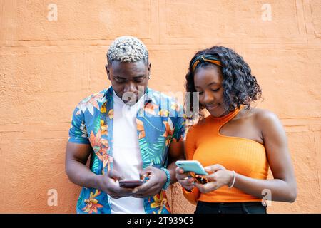 Giovane coppia nera con abiti colorati e casual appoggiati alla parete mentre entrambi usano il loro smartphone Foto Stock