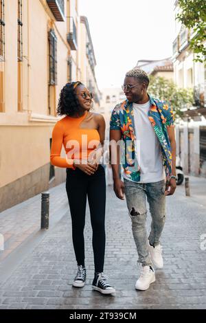 Giovane coppia nera che indossa occhiali da sole e abiti colorati che si guardano mentre cammina per la città in una giornata di sole Foto Stock