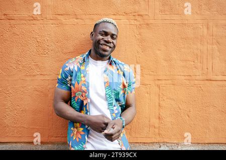 Ritratto di un bel giovane uomo nero che indossa abiti casual che guarda la fotocamera sorridere mentre si appoggia a una parete arancione Foto Stock