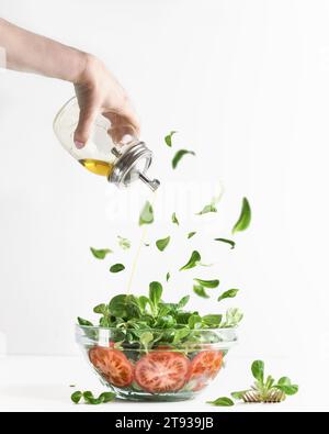 La donna versa a mano olio d'oliva sull'insalata verde in un recipiente di vetro con pomodori e foglie di lattuga volanti su sfondo bianco. Stile di vita sano. Insalata maki Foto Stock