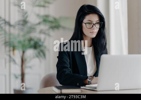 Donna professionale dedicata che lavora intensamente al suo laptop in un luminoso spazio ufficio con arredamento vegetale Foto Stock