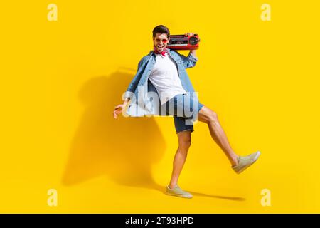 Foto a grandezza naturale di un ragazzo felicissimo che indossa una giacca in jeans con una passerella sul box portabagagli in uno spazio vuoto isolato su sfondo giallo Foto Stock