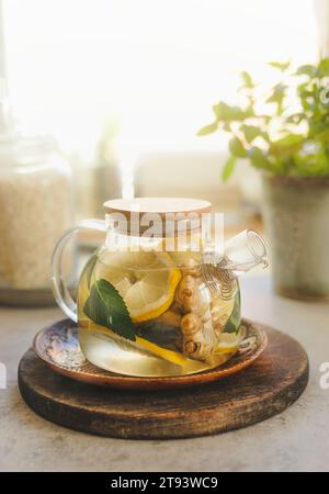 Teiera di vetro con limone, menta e zenzero su tagliere di legno sullo sfondo della finestra con luce naturale. Tè sano con vitamina c in tempo freddo. Fro Foto Stock