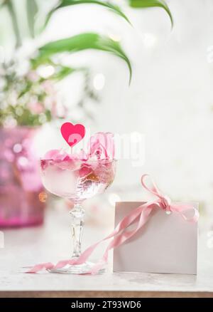 Bicchiere di champagne con petali rosa, cuore e carta regalo bianca bianca con nastro rosa su tavolo bianco su sfondo bokeh floreale. Festeggiamo valenti Foto Stock