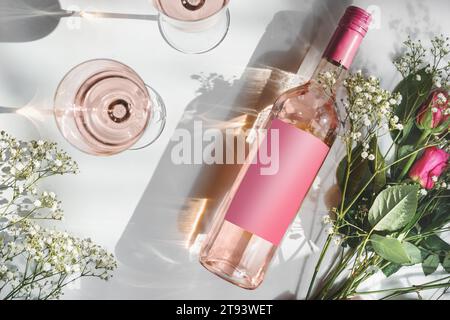 Bottiglia di vino rosa con etichetta vuota rosa, due bicchieri da vino e bouquet romantico di fiori su sfondo bianco con luce solare e ombra. Vino moderno Foto Stock