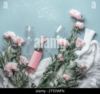 Bottiglia di vino rosato con verricelli su sfondo blu con maglione lavorato bianco e molti fiori rosa pallidi peonie. Vista dall'alto. Bellezza Foto Stock