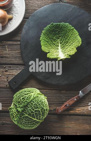 Cavolo crudo intero e foglia di cavolo sul tagliere nero su tavola rustica in legno da cucina con coltello. Preparazione del cibo con vegetab stagionale autunnale Foto Stock