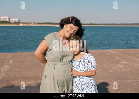 Una nonna con un abito verde abbraccia sua nipote con un abito a motivi sia in piedi in riva al mare con una costa lontana e edifici nel Foto Stock
