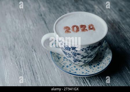 una tazza in ceramica bianca, con motivo floreale blu, con cappuccino e il numero 2024, come il nuovo anno, sulla sua schiuma di latte, su un tavolo in legno rustico grigio Foto Stock