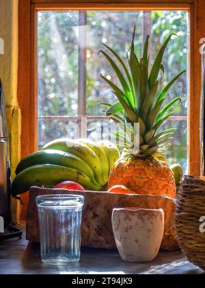 Un bicchiere e una tazza di argilla davanti a un vassoio di legno con banane, un ananas e alcune mele vicino a una finestra in una fattoria nel centro della Colombia. Foto Stock