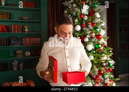 Giovane eccitato che apre il regalo di Natale a casa sua. Ritratto di un uomo felice ed emotivo che apre la confezione regalo di Natale. Foto Stock