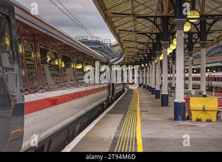 Una carrozza ferroviaria si trova su una piattaforma della stazione dove le luci di un tettuccio si riflettono sul treno. Un membro aspetta per una rampa. Foto Stock