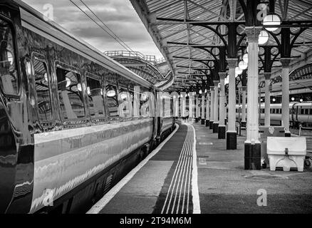 Una carrozza ferroviaria si trova su una piattaforma della stazione dove le luci di un tettuccio si riflettono sul treno. Un membro aspetta per una rampa. Foto Stock