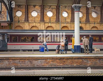 I passeggeri sono sul binario della stazione mentre il treno dietro di loro si prepara alla partenza. Tra di loro c'è una colonna del XIX secolo e un muro di mattoni oltre. Foto Stock