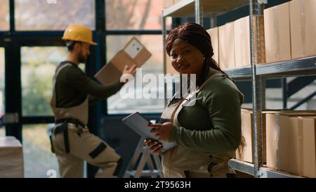 Ritratto di un gioioso responsabile della logistica in magazzino che tiene tablet, che controlla l'imballaggio mentre il tirocinante in background gestisce gli articoli confezionati in scatole di cartone pronte per la spedizione Foto Stock