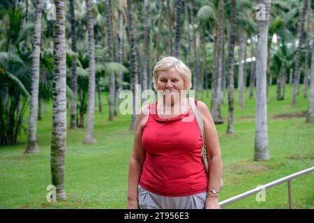 Una donna anziana sorridente si posa su uno sfondo tropicale verde brillante con palme. Foto Stock