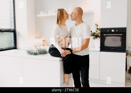 Felice coppia giovane aspetta il bambino in piedi insieme contro la finestra in casa, amorevole marito teneramente toccando la pancia di sua moglie incinta, spazio libero Foto Stock