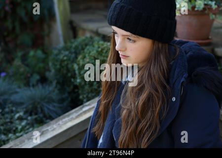 Ragazza seduta sul gradino che indossa un cappello di lana, un cappotto e una sciarpa che cerca di tenersi al caldo Foto Stock