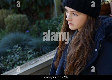 Ragazza seduta sul gradino che indossa un cappello di lana, un cappotto e una sciarpa che cerca di tenersi al caldo con le piante sullo sfondo Foto Stock