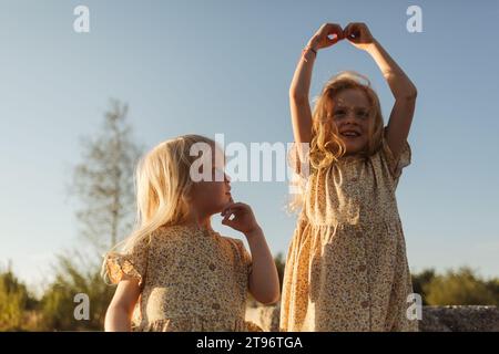 Dal basso bambina che guarda con curiosità la sorella maggiore che solleva le mani contro il cielo sfocato la sera indossando abiti coordinati Foto Stock