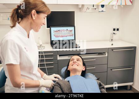 Vista laterale di una dentista adulta positiva in uniforme bianca sorridente mentre parla con la paziente sdraiata sulla sedia vicino a un computer portatile con la foto dei denti 3D sul display Foto Stock