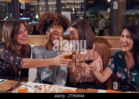 Gruppo di amiche femminili sorridenti e multietniche che brindano con occhiali da vista mentre si siedono seduti al tavolo del ristorante durante la celebrazione di un evento festivo Foto Stock