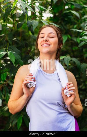 Giovane donna positiva in abiti casual con gli occhi chiusi sorridenti, contro le piante verdi e tenendo in mano un asciugamano da palestra intorno al collo Foto Stock