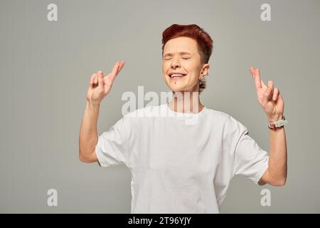 tesa persona bigender con t-shirt bianca che incrocia le dita per la fortuna in piedi con gli occhi chiusi sul grigio Foto Stock