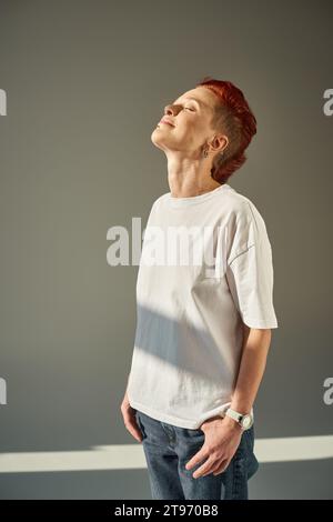 persona queer felice e unica con t-shirt bianca e jeans in piedi alla luce del sole su sfondo grigio Foto Stock