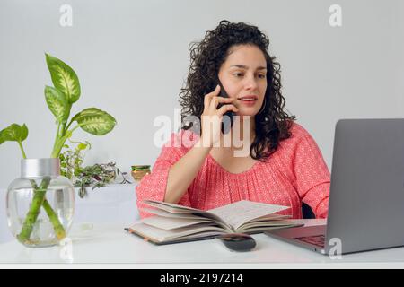 Giovane donna latina con arricciacapelli, vestita di rosa, seduta in ufficio a lavorare, a parlare al telefono e a usare un computer portatile con notebook aperto su tavolo e piante. Foto Stock