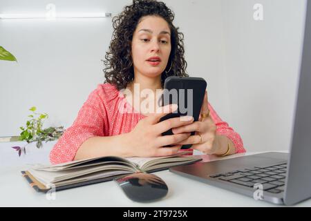 Giovane donna venezuelana latina, lavoratrice nel suo ufficio domestico seduta con il suo portatile sorpresa con la bocca aperta mentre leggeva messaggi telefonici e social media Foto Stock