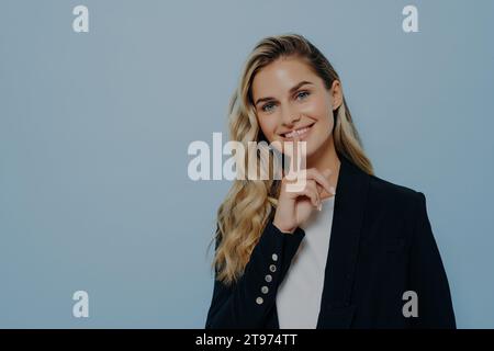 Donna elegante con blazer nero, dita a labbra, sorriso segreto, sfondo blu Foto Stock