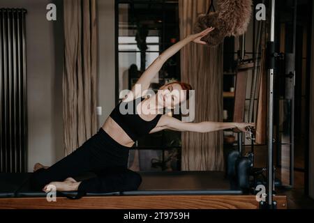Elegante rossa che esegue un tratto di Pilates laterale su un riformatore, mostrando grazia e flessibilità in un ambiente da studio sereno Foto Stock