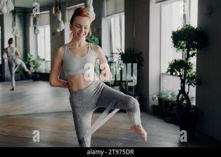 Gioiosa donna rossa in abbigliamento da palestra pratica con grazia gli esercizi della resistenza in uno spazio di allenamento illuminato dal sole e pieno di piante Foto Stock