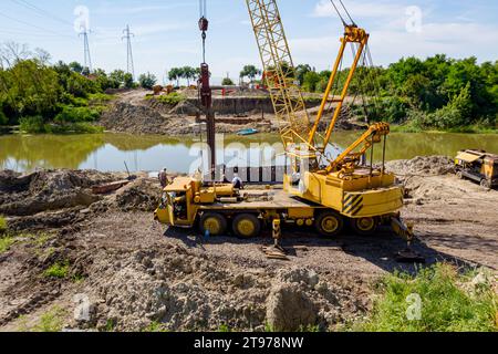 Gru mobile gialla obsoleta con martello pneumatico per l'installazione di pali metallici lungo la riva del fiume per la fondazione di ponti nel cantiere. Foto Stock