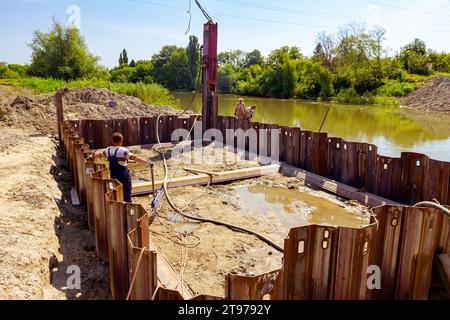 Il carrello gru mobile con martello pneumatico sta installando pali in metallo nelle fondamenta di ponti lungo la riva del fiume in cantiere. Il lavoratore gestisce con cra Foto Stock