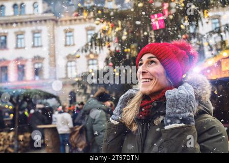 Donna gioiosa in berretto rosso che guarda la neve che cade al mercatino di Natale, con spazio per il copyspace per il tuo testo individuale. Foto Stock